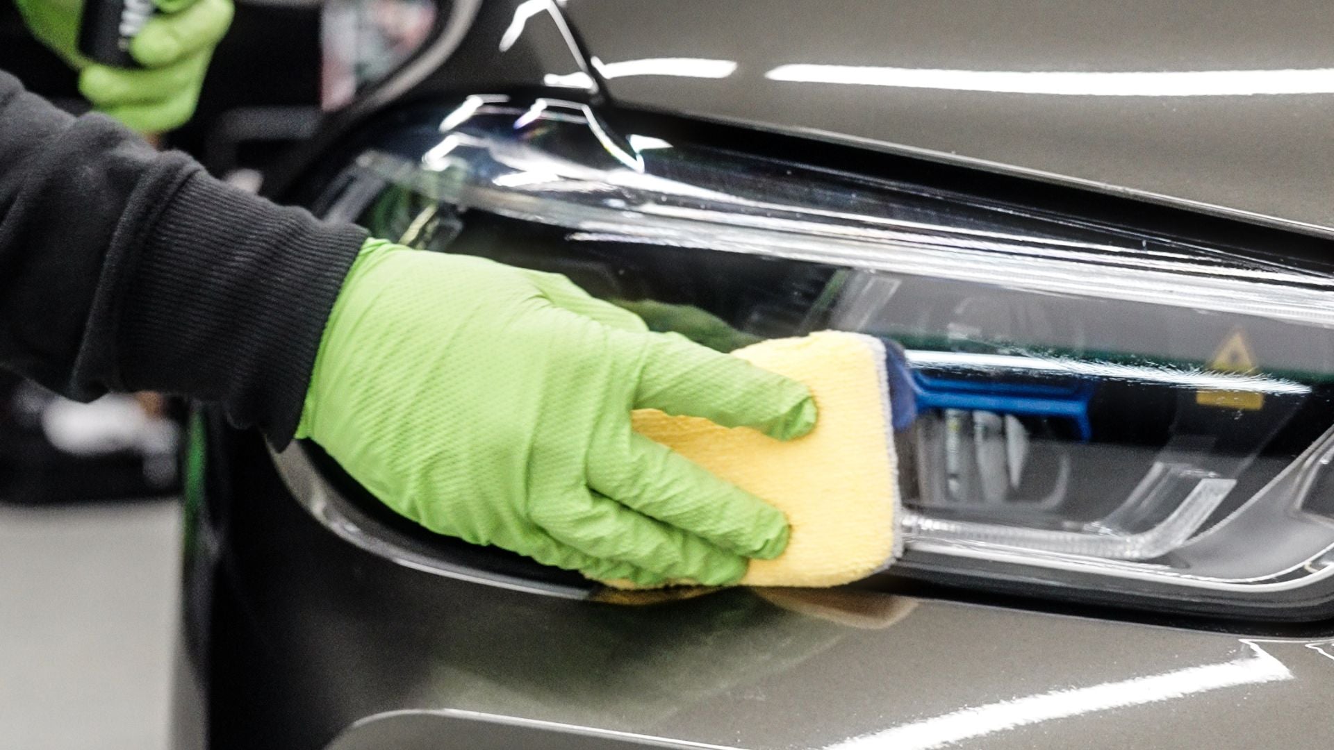 Detailer in green gloves applying ceramic coating to a car headlight with a microfiber applicator pad.